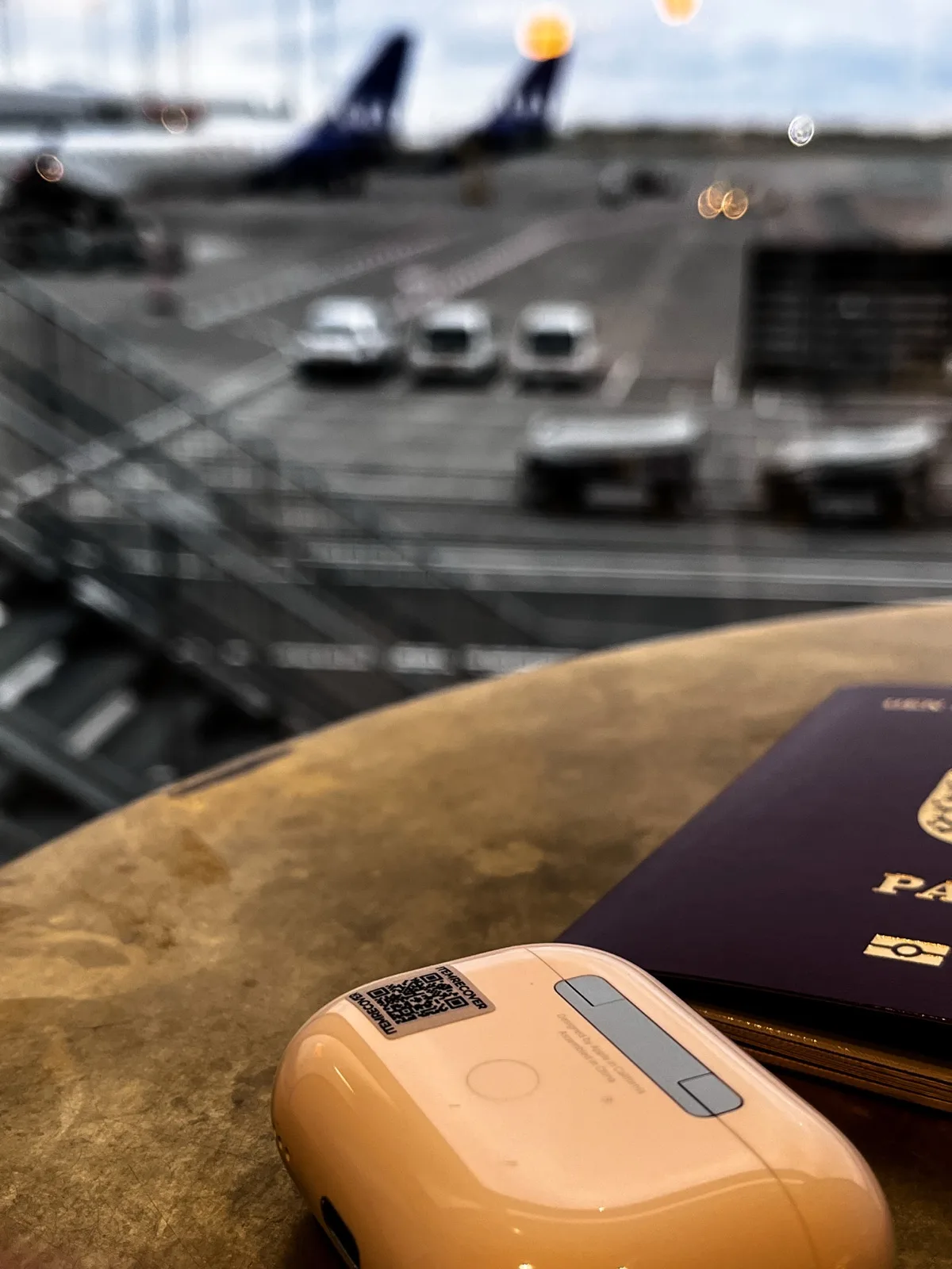 A passport, AirPods with an ItemRecover QR tag, and an airport runway visible through the lounge window